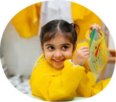 Child in yellow apron holding artwork during sensory workshop at Piccoli.