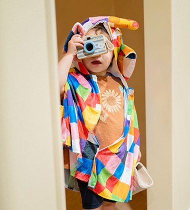 Child dressed in colorful costume pretending to take photos during creative role-play session at Piccoli.